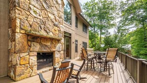 A wooden deck with a stone fireplace, outdoor seating, and a view of trees. - The Haven by Taylor Made Deep Creek (Oakland)