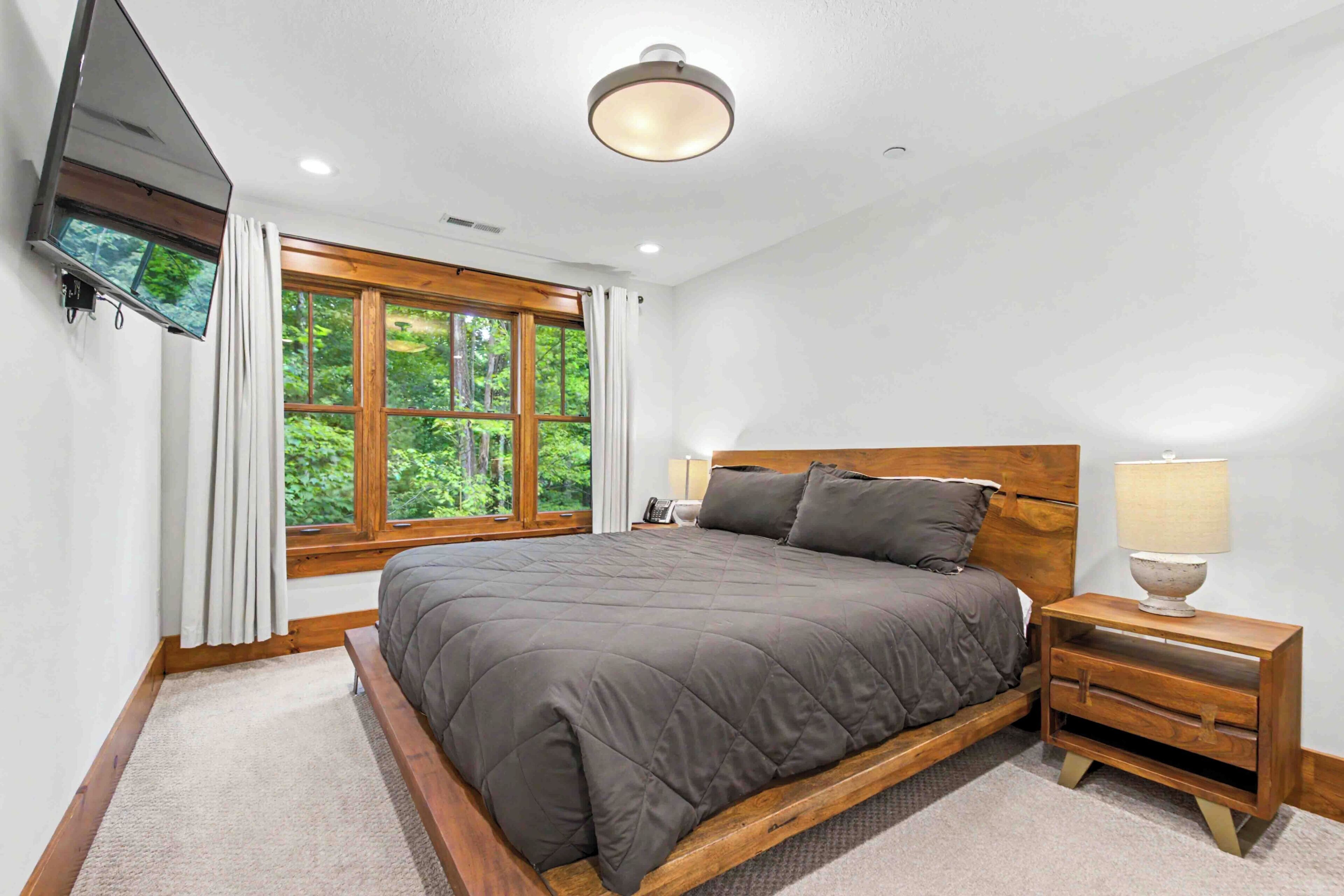 A bedroom with a large bed, a bedside table, a lamp, a TV mounted on the wall, and a window with curtains.