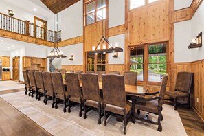 A spacious dining area with a long wooden table and upholstered chairs, a staircase with a wooden railing, and large windows offering views of the outdoors. - The Haven by Taylor Made Deep Creek (Oakland)