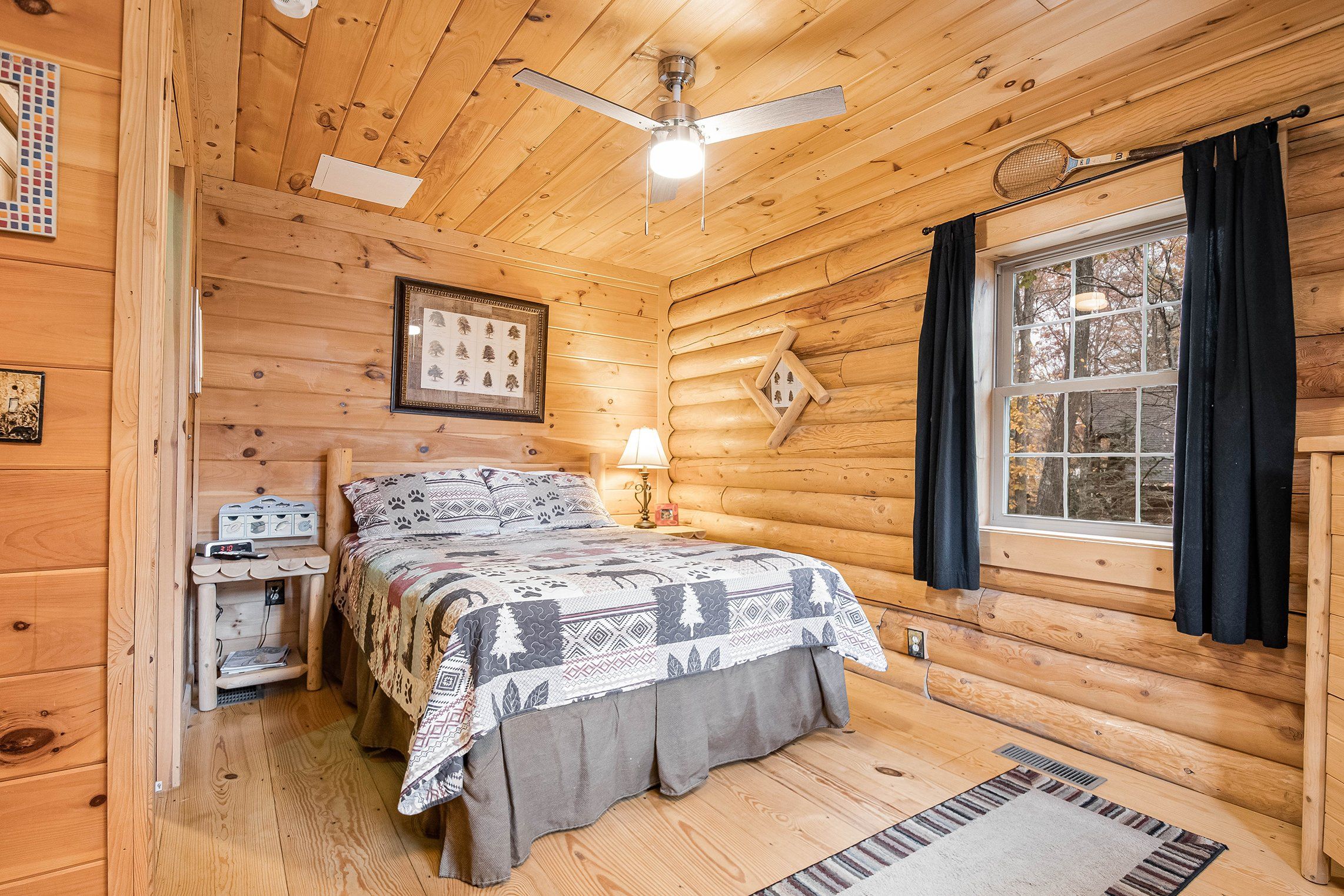 A log cabin bedroom with a bed, a ceiling fan, a window with curtains, and a wall-mounted shelf.
