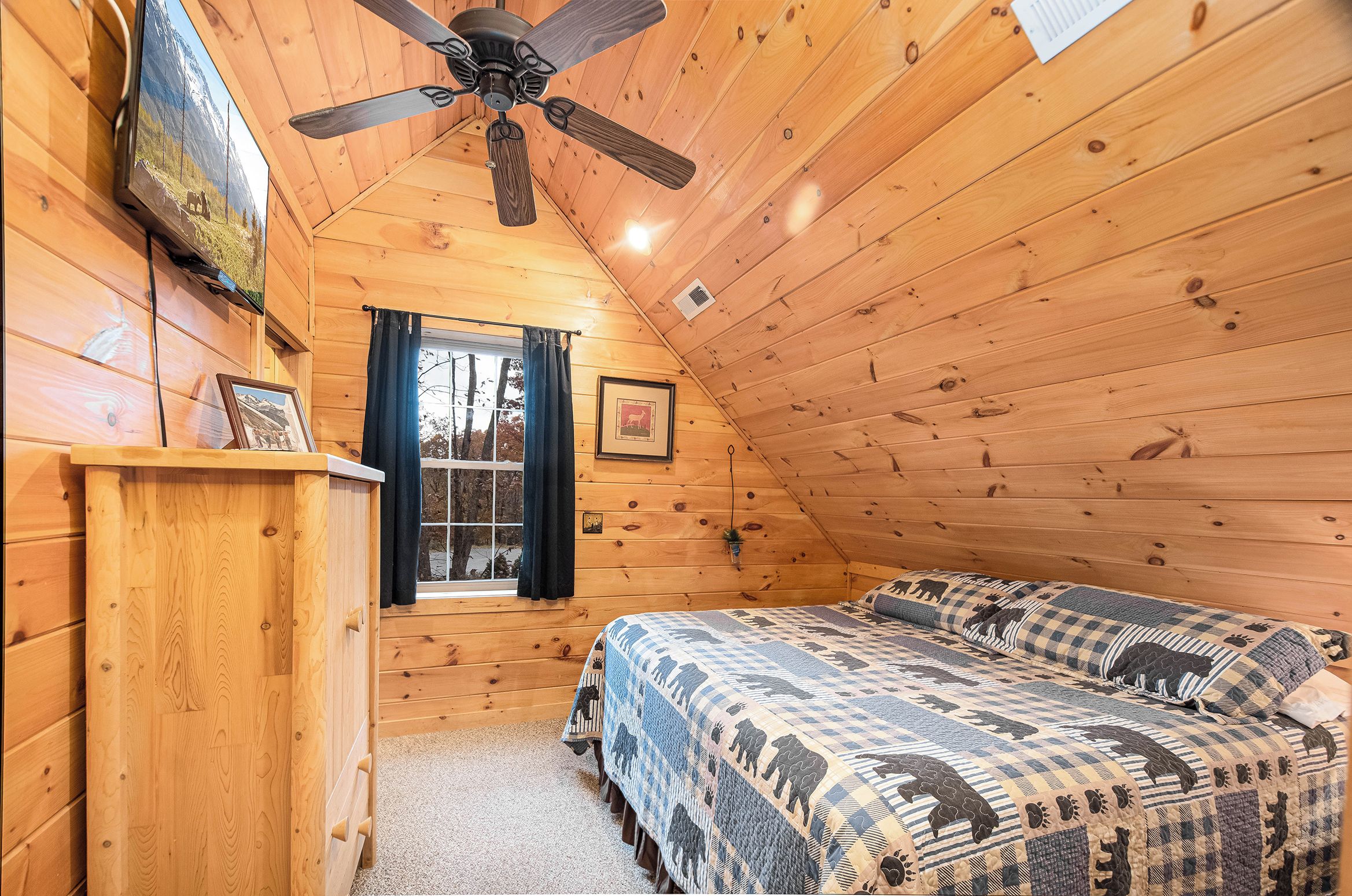 A cozy wooden cabin bedroom with a bed, a ceiling fan, a TV, and a window with curtains.
