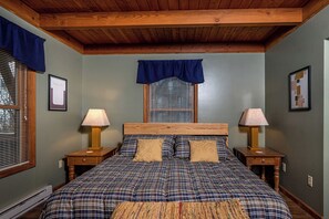 A bedroom with a wooden ceiling, a large bed with plaid bedding, two bedside tables with lamps, and a window with blinds. - The Perch by Taylor Made Deep Creek (Oakland)