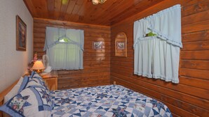 Une chambre dans une cabane en bois, avec un lit, deux fenêtres avec des rideaux, une table de chevet avec une lampe et un ventilateur fixé au mur.