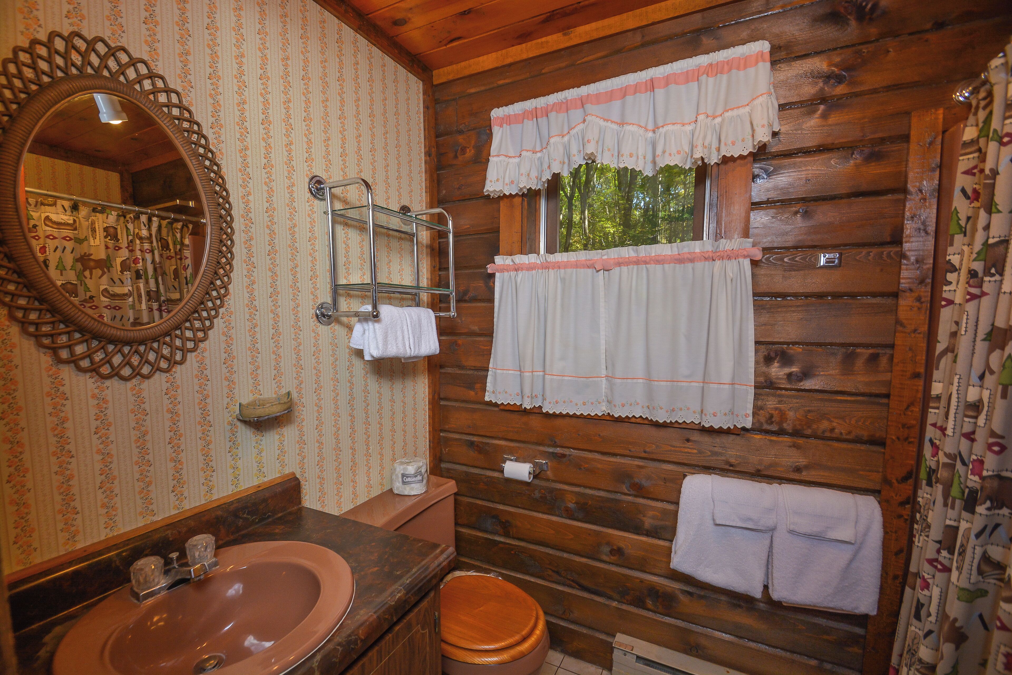 Un bagno in una cabina di legno con lavabo in legno, uno specchio, un water e una finestra con tende.
