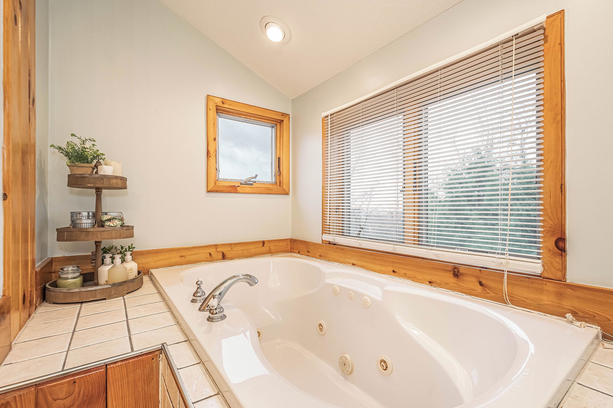 A bathroom with a large bathtub, wooden window blinds, a mirror, and a shelf with toiletries.