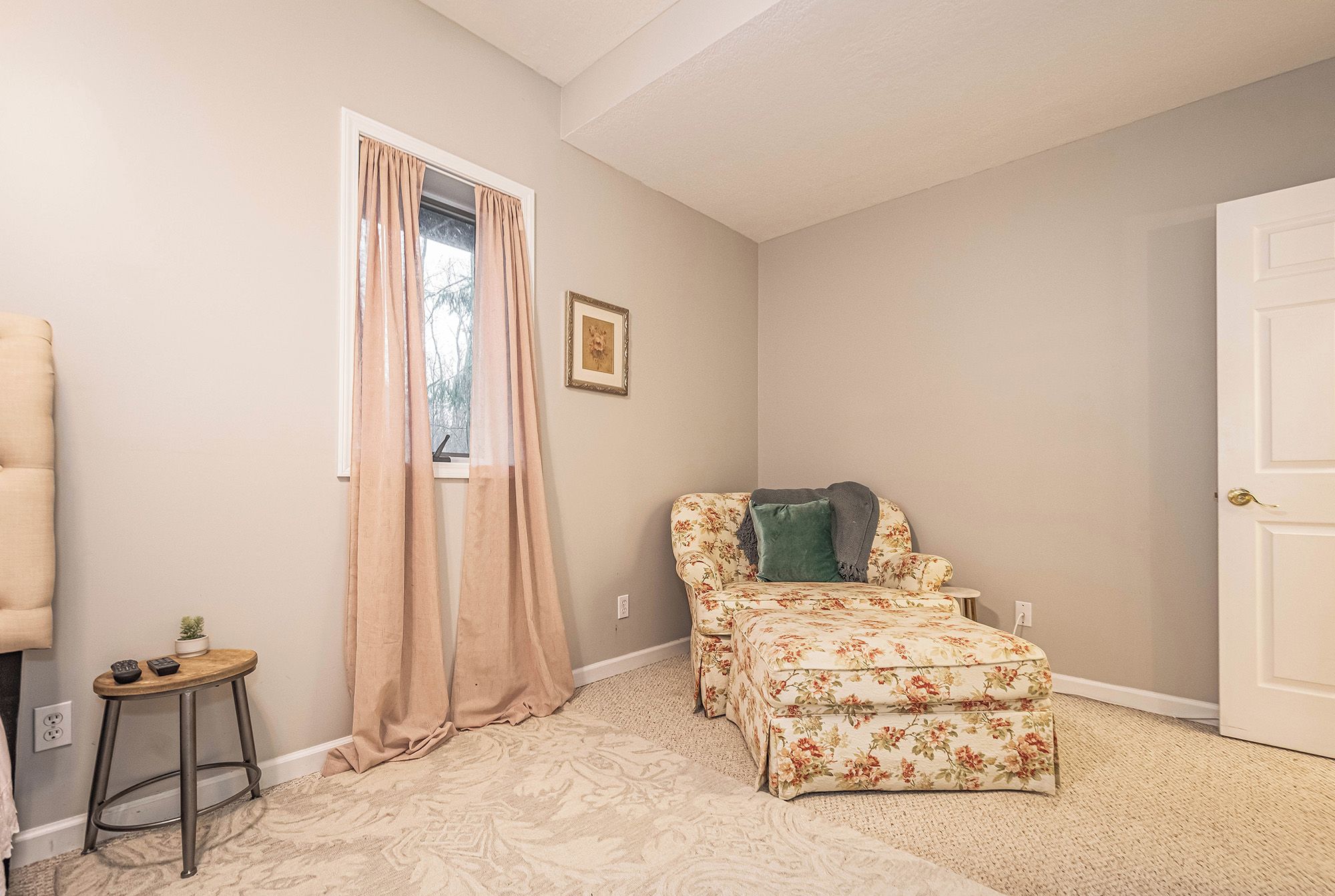 A room with a floral sofa, a matching ottoman, a small side table with a plant, and a window with sheer curtains.