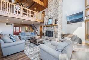 A spacious living area with a stone fireplace, a balcony, and a large TV mounted on the wall. - Under the Stars by Taylor Made Deep Creek (Oakland)