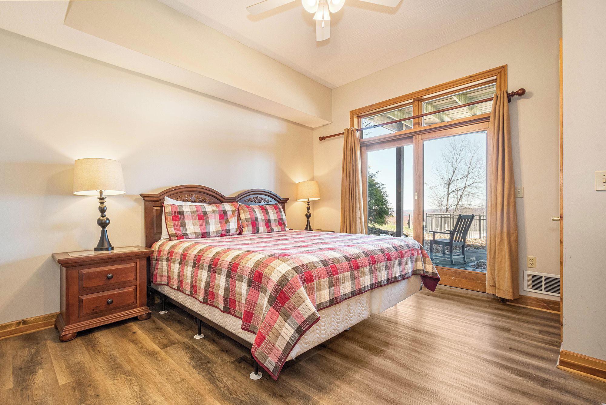 Un dormitorio con cama de madera, dos mesitas de noche con lámparas, un ventanal con cortinas y vistas al exterior.