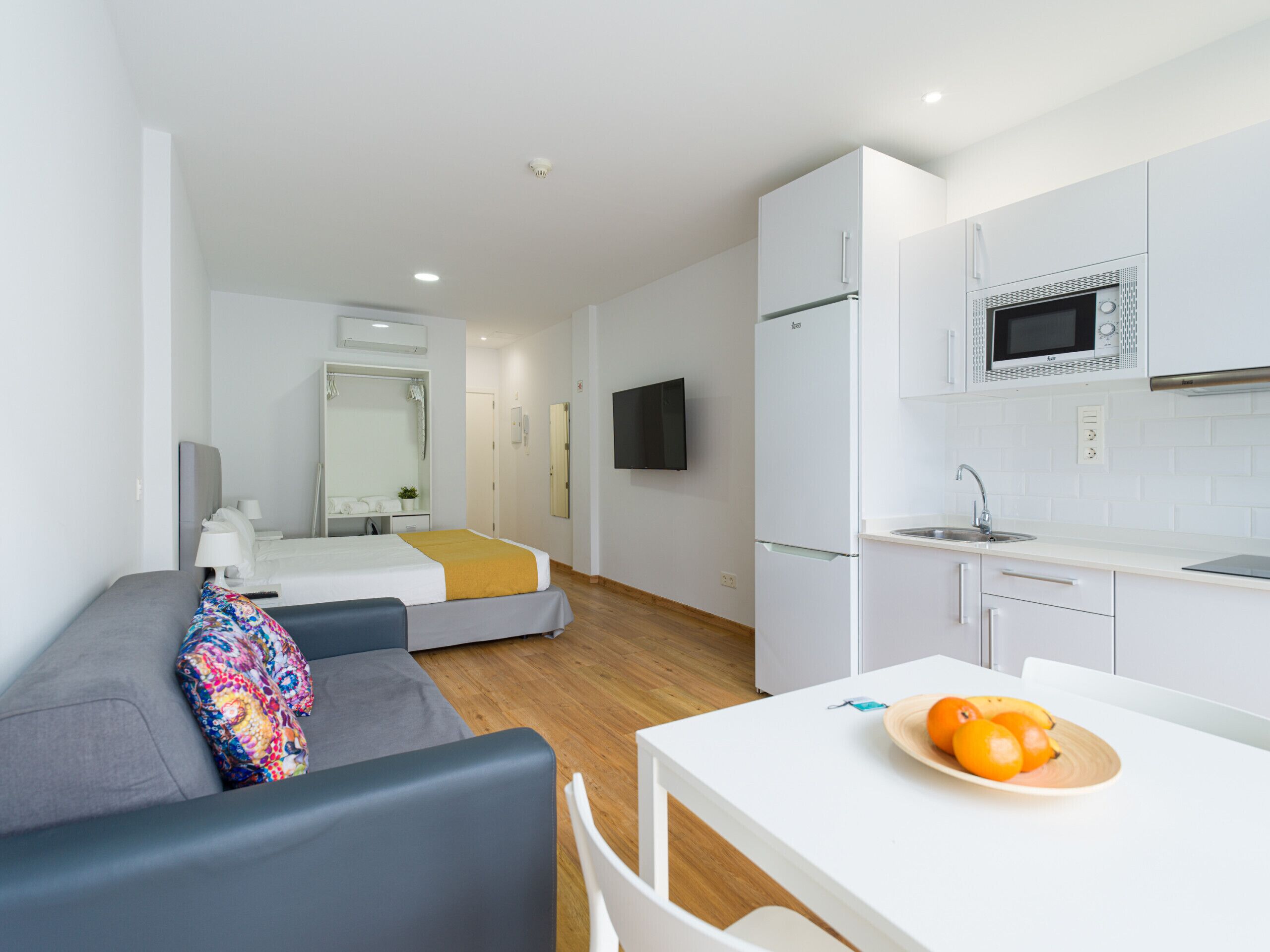 A modern hotel room with a bed, a sofa, a small table with fruit, a microwave, and a TV.
