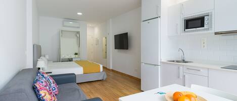 A modern hotel room with a bed, a sofa, a small table with fruit, a microwave, and a TV.