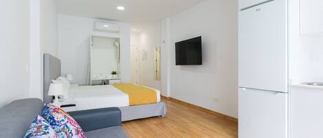 Una habitación de hotel moderna con un sofá gris, una cama con manta amarilla, un armario blanco, un televisor de pantalla plana montado en la pared y suelo de madera.