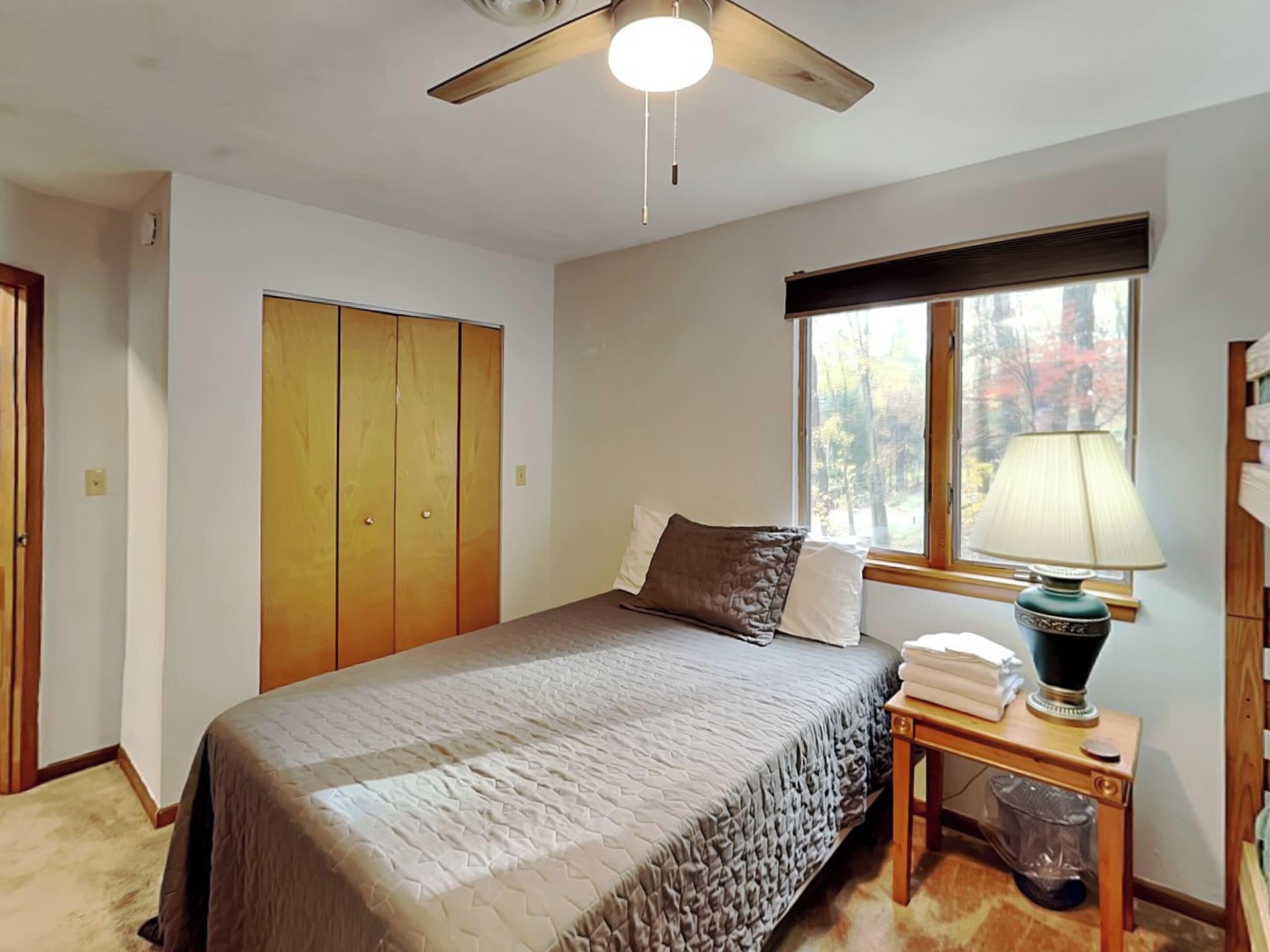 A bedroom with a bed, a ceiling fan, a window with a view of trees, and a bedside table with a lamp.