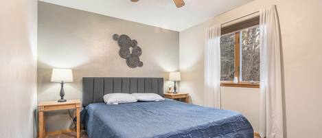 A bedroom with a bed, two bedside tables with lamps, a ceiling fan, a window with curtains, and a wall decoration.
