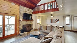 A spacious living room with a stone fireplace, a large sofa, a television mounted on the wall, and a balcony with outdoor furniture.