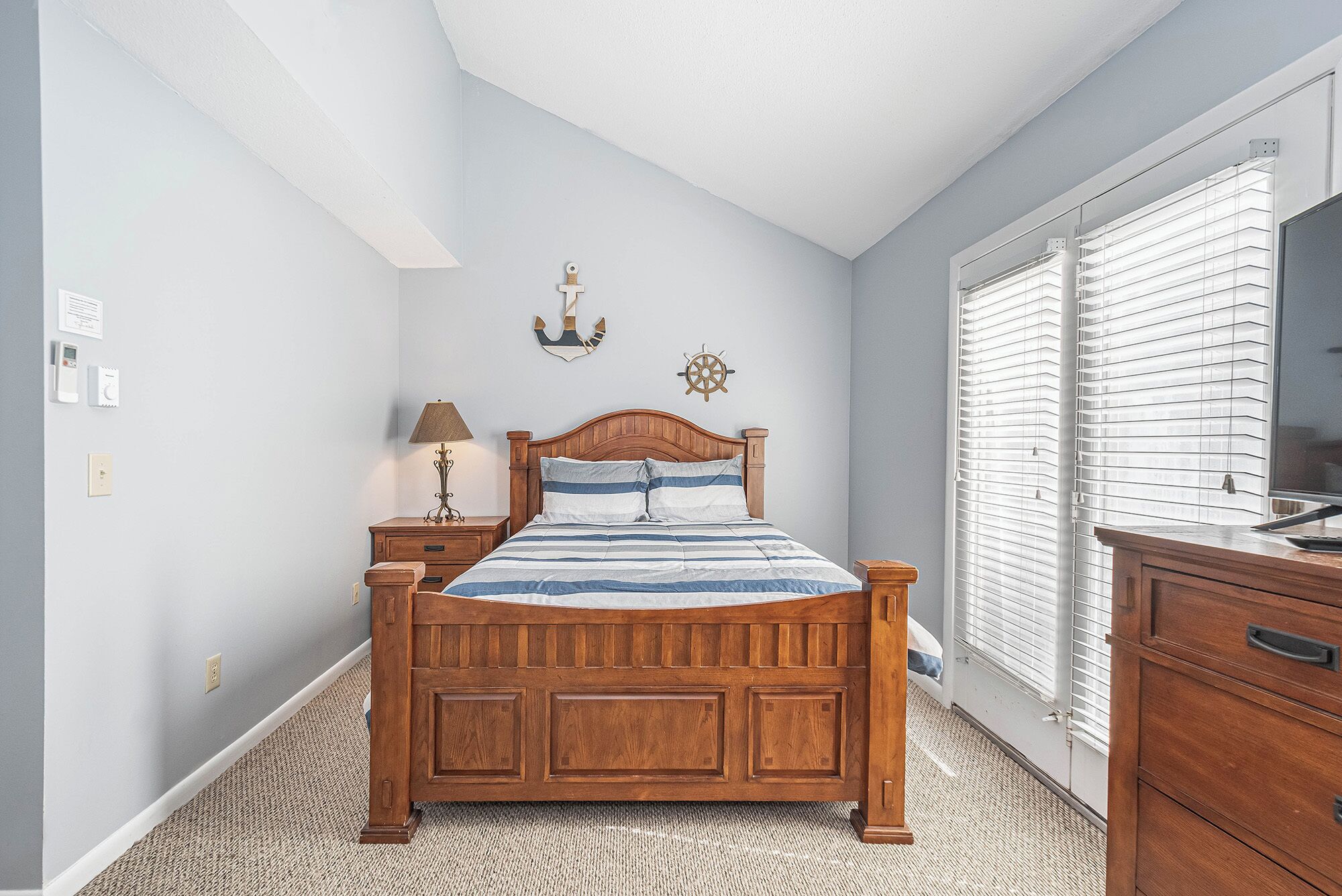 A bedroom with a wooden bed, a nightstand, a lamp, a wall clock, and a window with blinds.