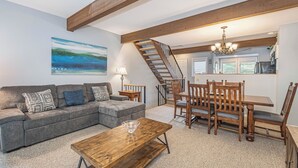 A living room with a gray sectional sofa, a wooden coffee table, and a dining area with wooden chairs and a table. A staircase with a wooden railing leads to an upper level. - Villages of Wisp 08 Winding Way by Taylor Made Deep Creek (McHenry)