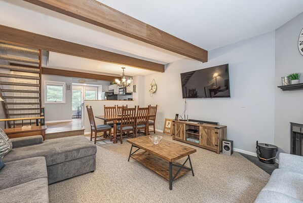 A spacious living room with a wooden dining table, grey sofa, and a staircase leading to a kitchen.