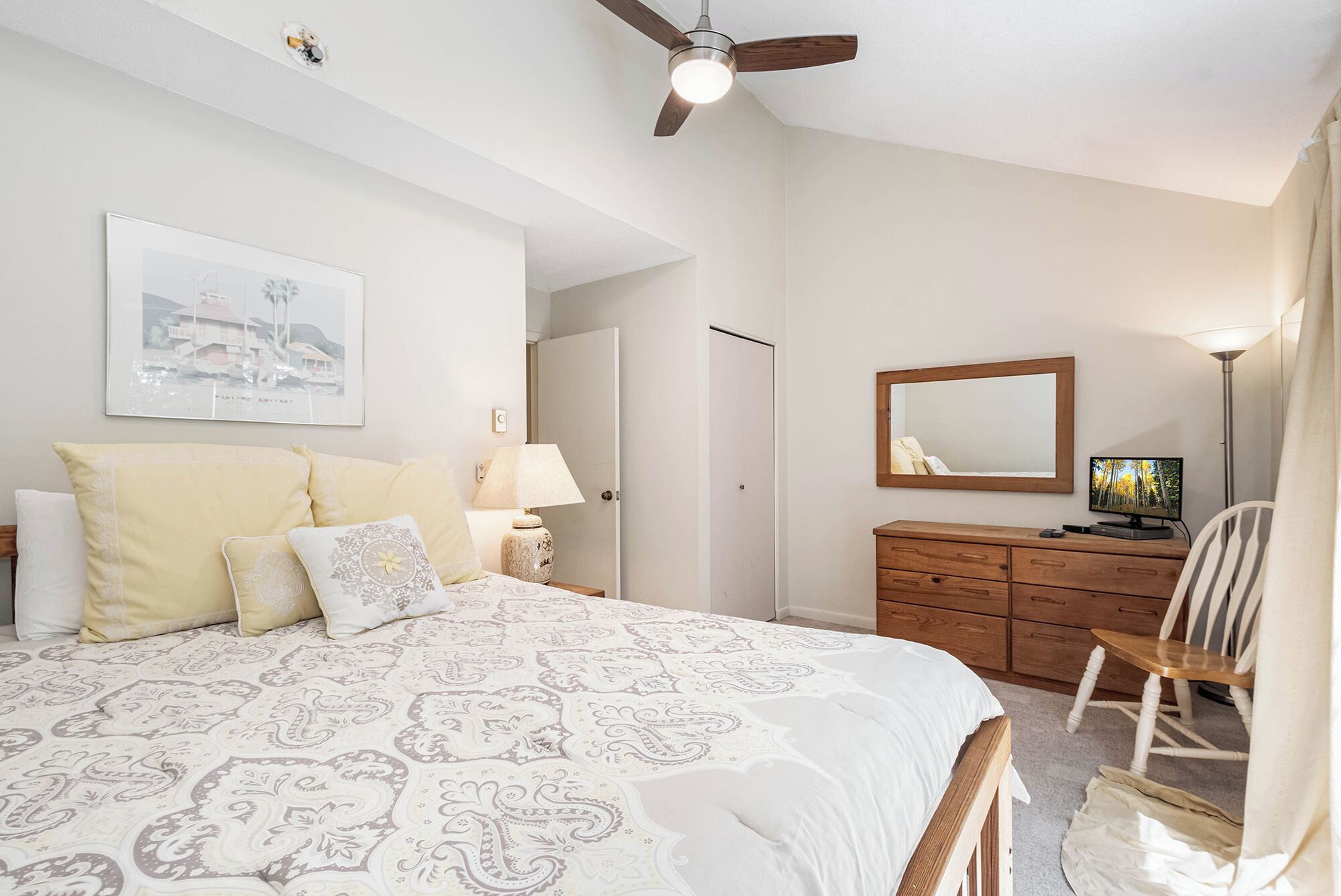 Un dormitorio con cama, cómoda, silla, ventilador de techo, espejo y televisor.
