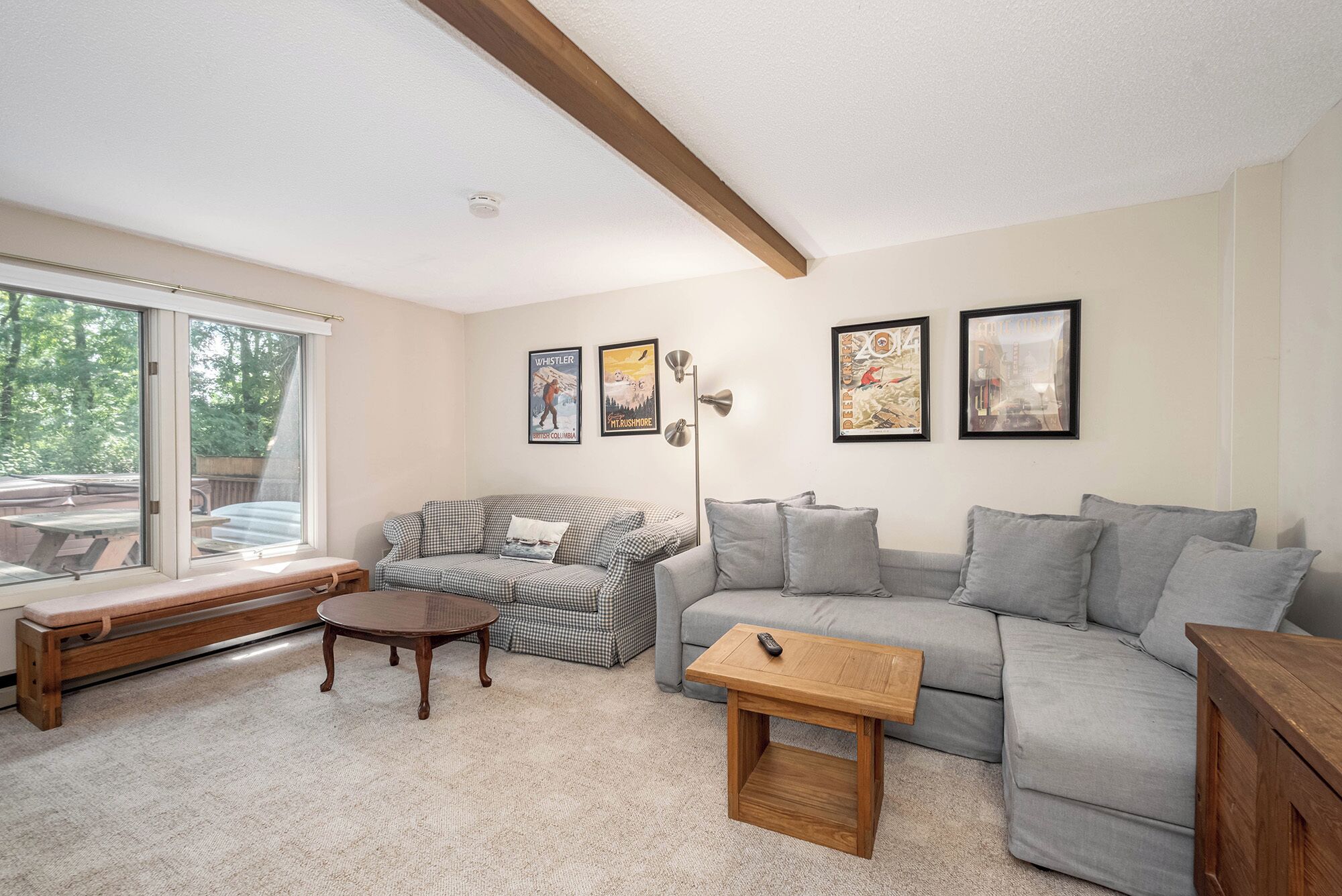 A living room with a checkered sofa, a grey sectional sofa, a wooden coffee table, and a bench. There are framed pictures on the wall and a window with a view of trees.