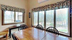 Un comedor con mesa y sillas de madera, ventanas grandes con cortinas y un equipo de aire acondicionado en la pared.