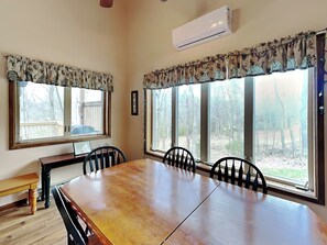 A dining room with a wooden table and chairs, large windows with curtains, and an air conditioning unit on the wall. - Wake at the Lake by Taylor Made Deep Creek (Swanton)