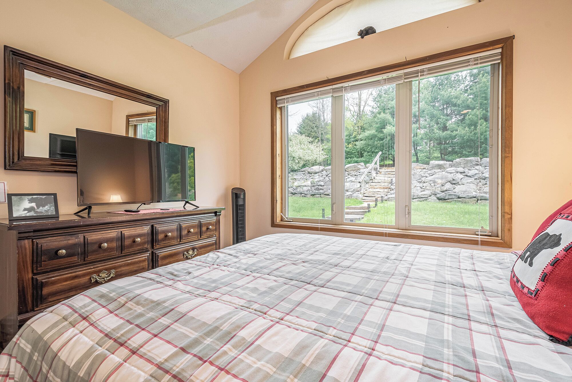 A bedroom with a large bed, a dresser, a TV, and a window with a view of a stone wall and greenery.