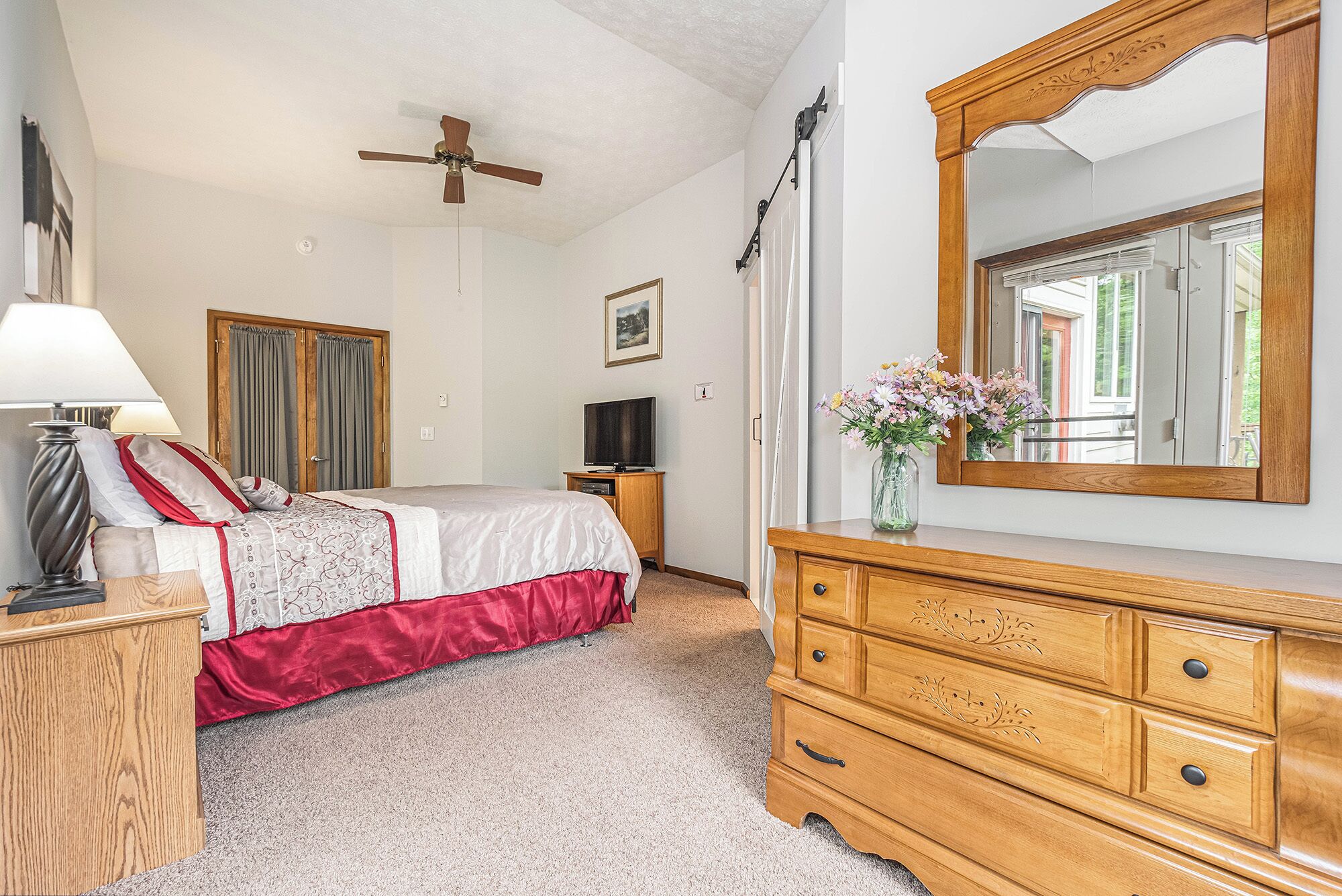 Une chambre à coucher comprenant un lit, une commode en bois, un miroir, un ventilateur de plafond et un téléviseur.
