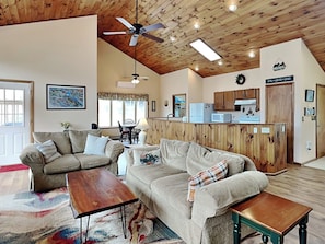 A cozy living room with a sofa, two coffee tables, and a ceiling fan. A kitchen with wooden cabinets and appliances is visible in the background. - Wake at the Lake by Taylor Made Deep Creek (Swanton)