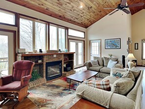 A spacious living room with a fireplace, a comfortable sofa set, a wooden ceiling, and large windows. - Wake at the Lake by Taylor Made Deep Creek (Swanton)