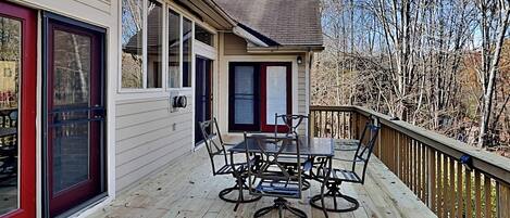 A wooden deck with a table and chairs, a sliding glass door, and a view of trees and mountains.