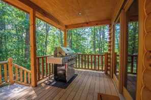 Eine überdachte Holzterrasse mit einem Grill, umgeben von einem Holzgeländer und mit Blick auf ein bewaldetes Gebiet.