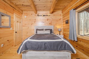 Ein Schlafzimmer einer Holzhütte mit einem Bett, einem Nachttisch, einer Lampe, einem Fenster mit Vorhängen und einem gerahmten Bild an der Wand.