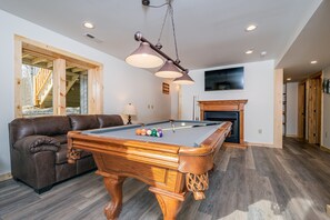 Ein Billardtisch mit Kugeln, eine Couch und ein Kamin in einem Raum mit Holzboden und einem an der Wand montierten Fernseher.