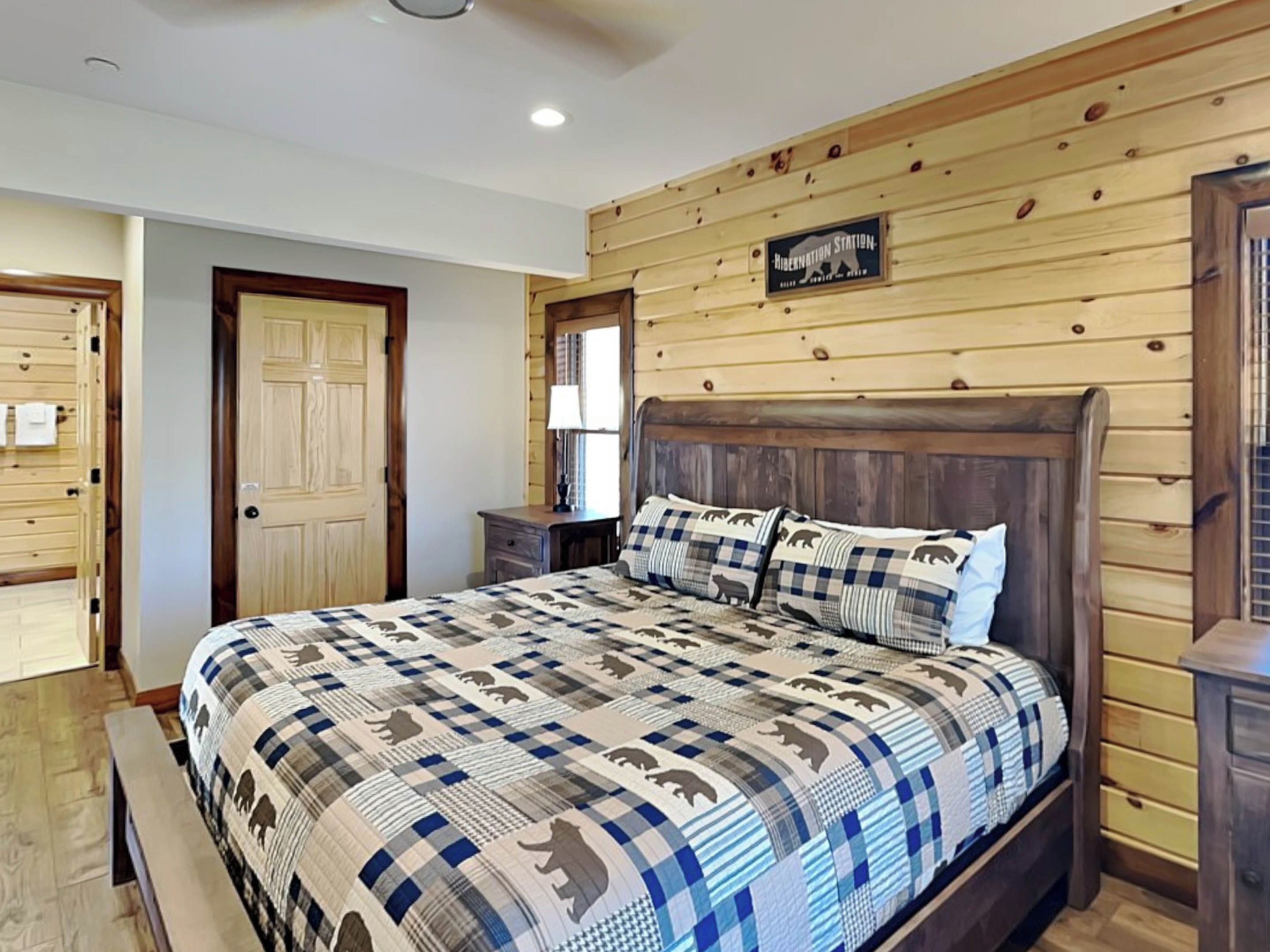 Quarto com painel de parede em madeira, uma cama grande com colcha estampada, uma mesinha de cabeceira e uma porta de madeira.