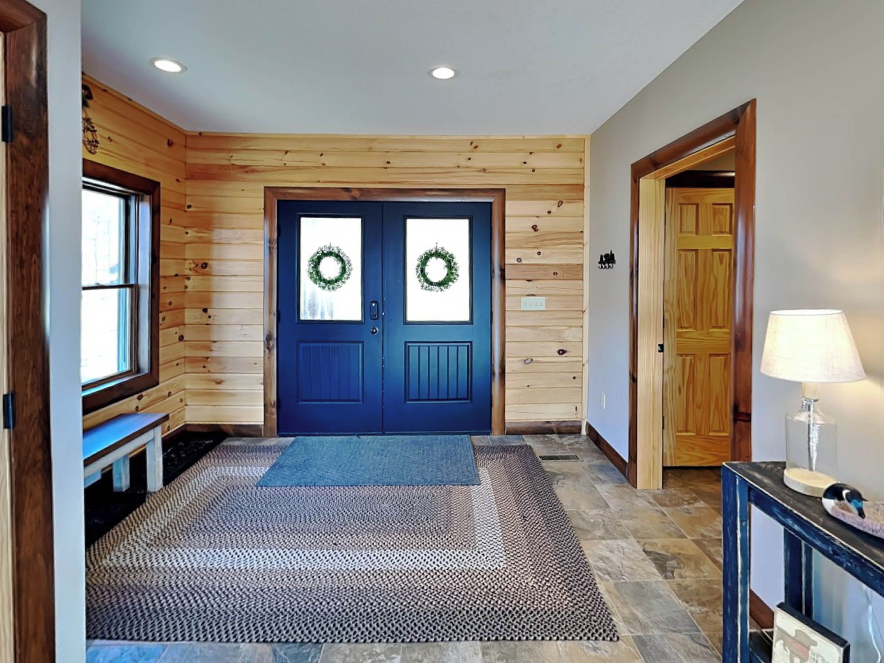 Interior de madera con una puerta azul, un felpudo y una lámpara sobre una mesa.