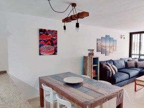 Una sala de estar con un sofá azul, una mesa de comedor de madera con sillas y una lámpara fijada a la pared.