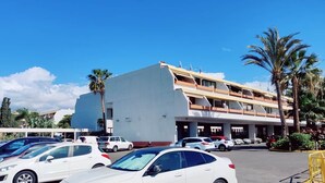 Un hôtel à plusieurs étages, avec un parking rempli de voitures, un ciel bleu azur et des palmiers.