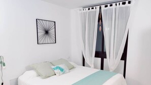 Un lit bien fait, recouvert d’une couvre-lit blanche et turquoise, agrémenté d’une œuvre d’art abstraite noire au mur, et d’un rideau blanc devant la fenêtre.