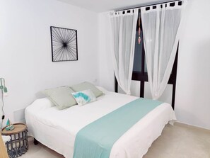 Una cama bien hecha con una colcha blanca y azul turquesa, una obra de arte abstracta negra en la pared y una cortina blanca sobre una ventana.