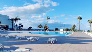 Un espace aménagé au bord de la piscine, avec des chaises longues, des palmiers et un ciel dégagé.