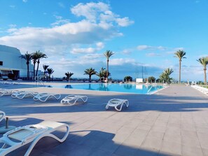 Un área junto a la piscina con sillas de descanso, palmeras y cielo despejado.