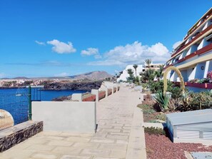A coastal hotel walkway with a view of the sea and mountains, a pool area, and a building with balconies.