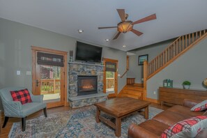 A living room with a stone fireplace, a ceiling fan, and a staircase. - Wine Down by Taylor Made Deep Creek (Oakland)