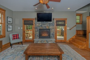 A living room with a stone fireplace, a flat-screen TV, and a ceiling fan. - Wine Down by Taylor Made Deep Creek (Oakland)