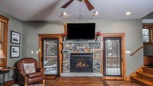 A living room with a stone fireplace, a leather armchair, a television, and a staircase. - Wine Down by Taylor Made Deep Creek (Oakland)