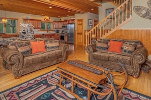 A spacious living room with two brown leather sofas, a wooden coffee table, and a patterned rug. A kitchen with wooden cabinets and a refrigerator is visible in the background. - Wisp N Around by Taylor Made Deep Creek (McHenry)