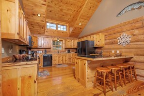 A wooden cabin interior with a kitchen featuring wooden cabinets, a refrigerator, a sink, and a bar area with stools. - Wispy Business by Taylor Made Deep Creek (McHenry)