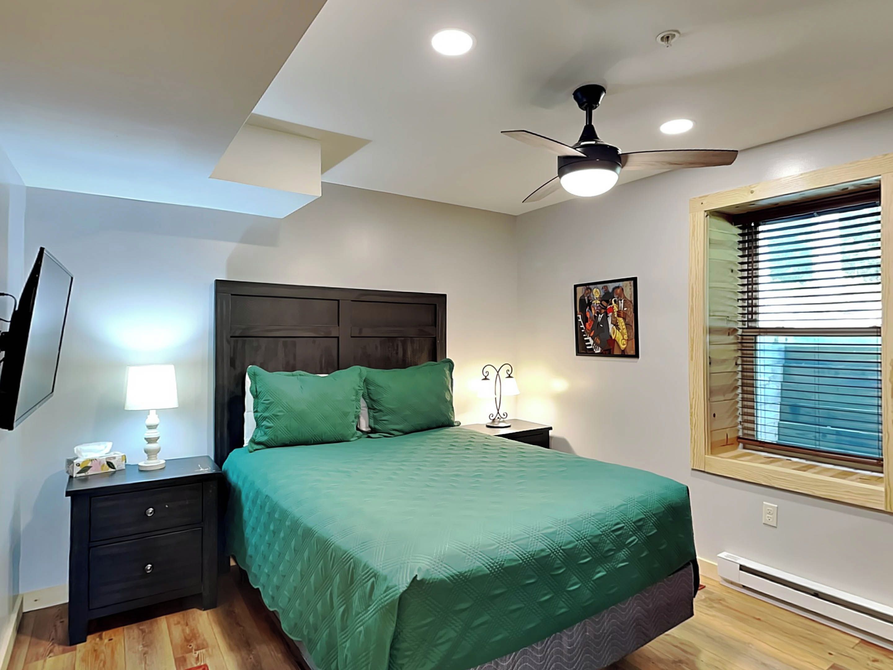 Une chambre à coucher comprenant un lit, une table de chevet, une lampe, un ventilateur de plafond, un tableau et une fenêtre avec des stores.
