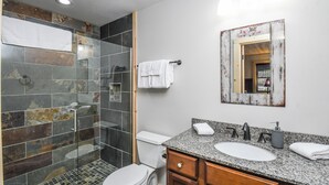 A bathroom with a glass-enclosed shower, a white toilet, a granite countertop sink, and a wooden vanity. - Woods-stock by Taylor Made Deep Creek (McHenry)