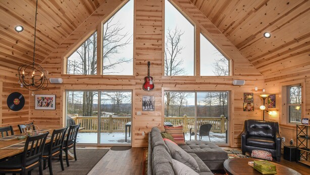 A log cabin interior with a dining area, a large sofa, a leather armchair, and a view through the windows. - Woods-stock by Taylor Made Deep Creek (McHenry)
